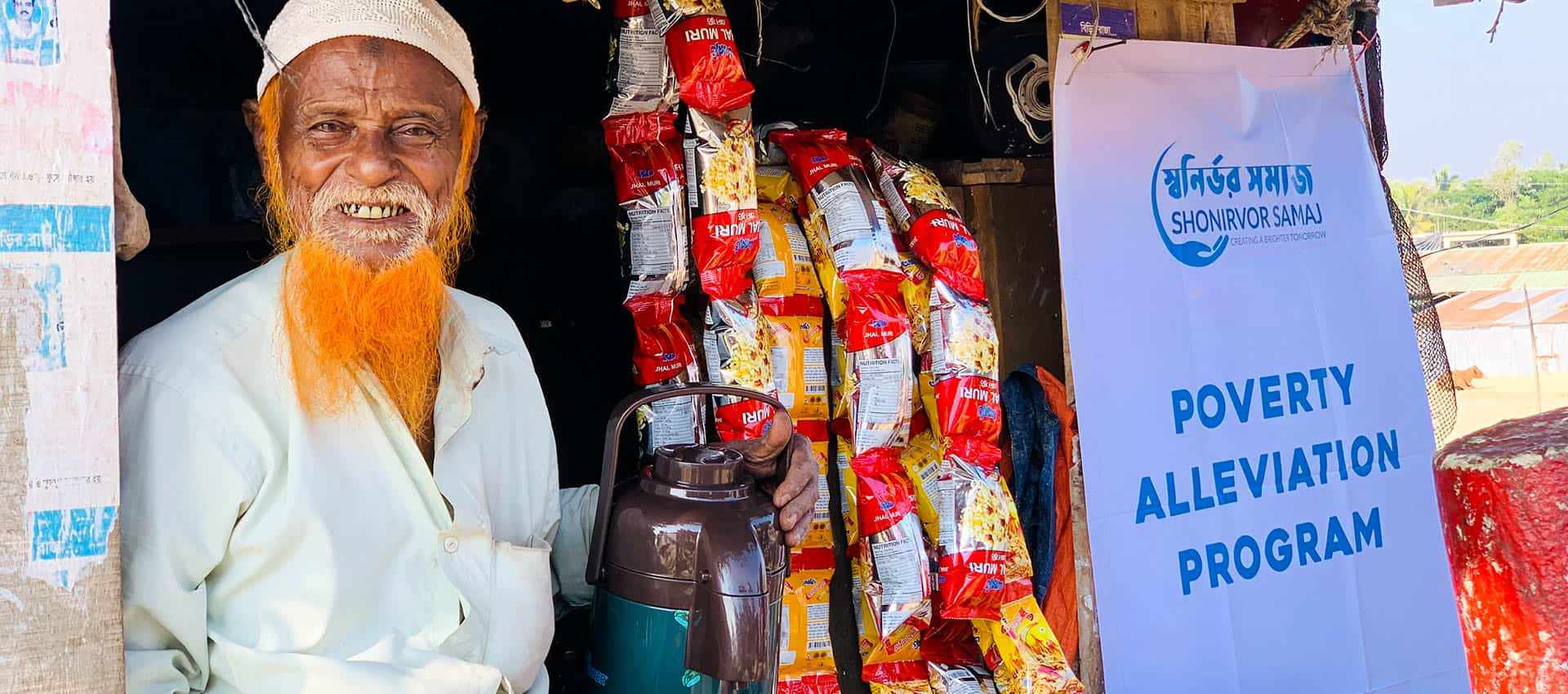 A smiling elderly man beside a Shonirvor Samaj poverty alleviation programme banner