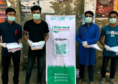 Four masked volunteers holding leaflets beside a Shonirvor Samaj banner with a QR code
