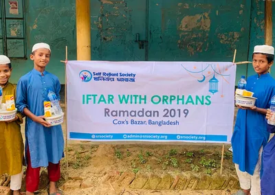 Boys holding iftar packs beside an Iftar with Orphans, Ramadan 2019 banner