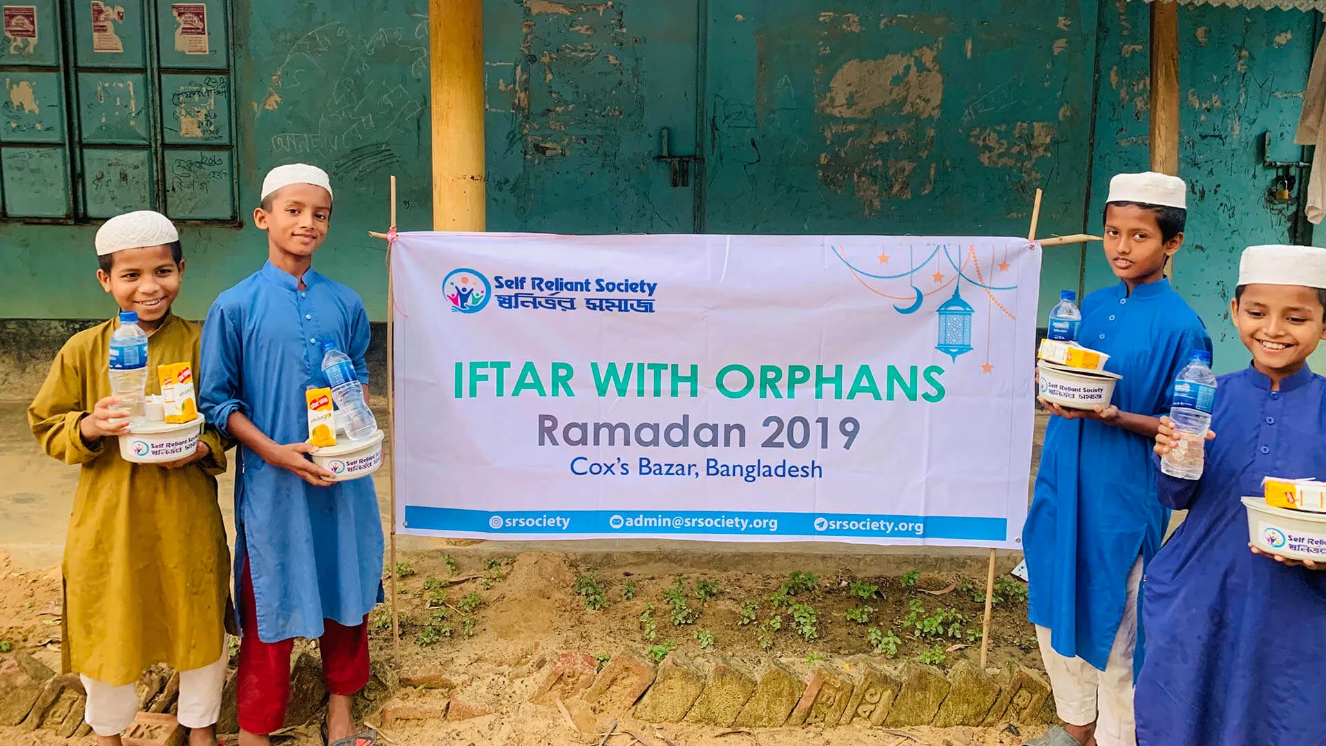Children holding iftar food packs beside an “Iftar with Orphans, Ramadan 2019” banner